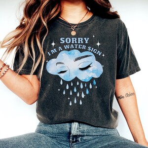 May include: A dark gray t-shirt with a blue watercolor cloud with eyes closed and raindrops falling. The text on the shirt reads "Sorry I'm a water sign".