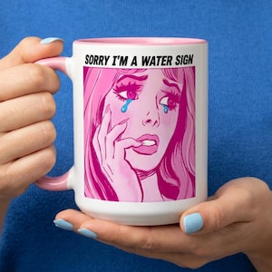 May include: White ceramic mug with a pink interior and handle. The mug features a pop art style illustration of a crying woman with pink hair and the text "SORRY I'M A WATER SIGN".