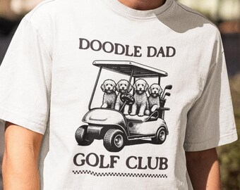 May include: A light grey t-shirt with the text "DOODLE DAD GOLF CLUB" printed in black. The graphic shows four doodle dogs in a golf cart. The person is wearing black sunglasses and a black cap.