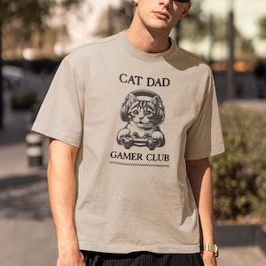 May include: A beige t-shirt with the text "CAT DAD GAMER CLUB" and a graphic of a cat wearing headphones and holding a game controller. The shirt is short-sleeved.