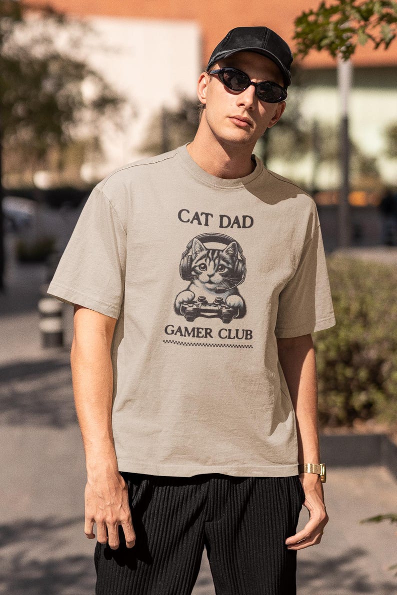 May include: A beige t-shirt with the text "CAT DAD GAMER CLUB" and a graphic of a cat wearing headphones and holding a game controller. The shirt is short-sleeved.