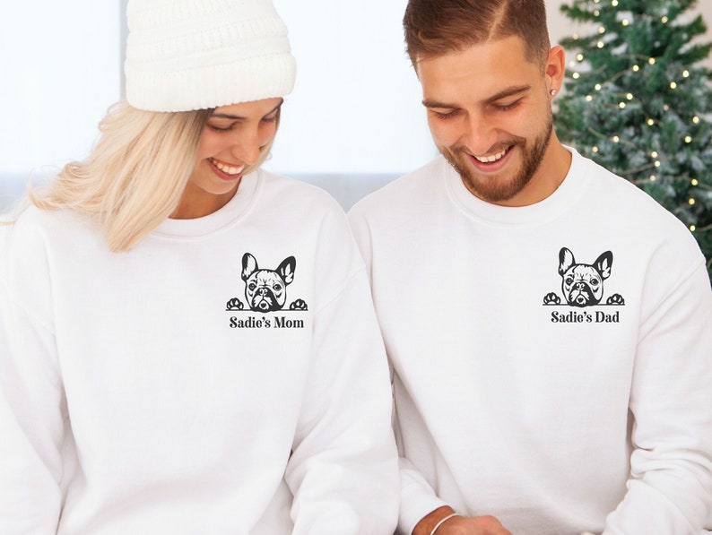May include: Two white sweatshirts with a black French bulldog peeking over the top. The sweatshirt on the left says "Sadie's Mom" and the sweatshirt on the right says "Sadie's Dad".