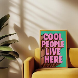 May include: A framed art print with the text "COOL PEOPLE LIVE HERE" in pink and orange, set against a teal background. The print is displayed on a yellow sofa, with a potted plant in the corner.