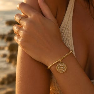 May include: Gold beaded bracelet with a round pendant featuring a zodiac symbol. The bracelet is worn on a wrist, and the pendant has a detailed design. Several gold rings are also visible on the fingers.