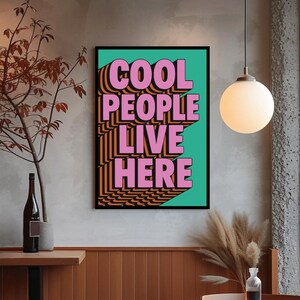 May include: A framed poster with the text "COOL PEOPLE LIVE HERE" in pink, orange, and black. The text is set against a teal background. The poster is in a room with a neutral color palette, a pendant light, and a wooden table.