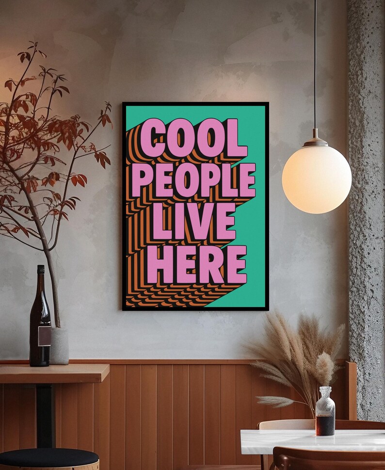 May include: A framed poster with the text "COOL PEOPLE LIVE HERE" in pink, orange, and black. The text is set against a teal background. The poster is in a room with a neutral color palette, a pendant light, and a wooden table.
