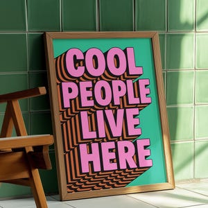 May include: A framed art print with the text "COOL PEOPLE LIVE HERE" in pink, orange, and black. The artwork is set against a teal background and framed in natural wood. The print is leaning against a green tiled wall.
