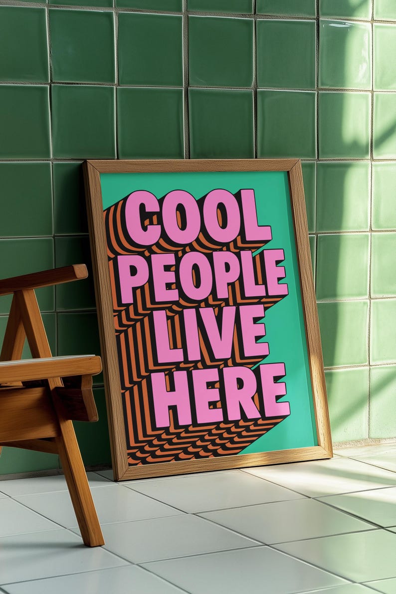 May include: A framed art print with the text "COOL PEOPLE LIVE HERE" in pink, orange, and black. The artwork is set against a teal background and framed in natural wood. The print is leaning against a green tiled wall.