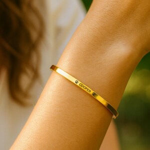 May include: A gold-colored cuff bracelet with the name "Cooper" and paw print symbols engraved on it. The bracelet is worn on a wrist, and the background is blurred.