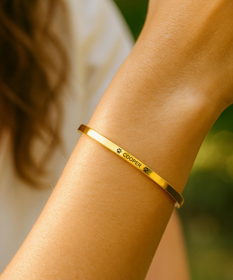 May include: A gold-colored cuff bracelet with the name "Cooper" and paw print symbols engraved on it. The bracelet is worn on a wrist, and the background is blurred.