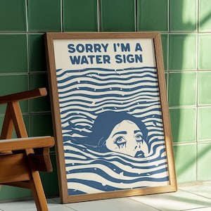 May include: A framed art print with the text "SORRY I'M A WATER SIGN" above a blue illustration of a person submerged in water. The artwork has a retro aesthetic, with a wooden frame and a white background.