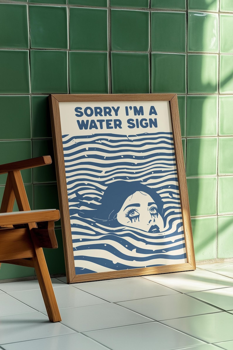 May include: A framed art print with the text "SORRY I'M A WATER SIGN" above a blue illustration of a person submerged in water. The artwork has a retro aesthetic, with a wooden frame and a white background.