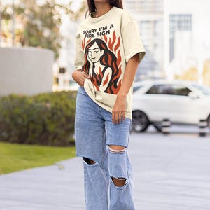 Puede incluir: Camiseta color crema con el texto "SORRY I'M A FIRE SIGN" sobre un gr&aacute;fico de una mujer con pelo rojo y llamas. La persona lleva gafas de sol y vaqueros rotos de color azul claro. La camiseta es una prenda informal.