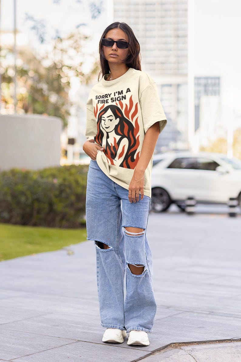 Puede incluir: Camiseta color crema con el texto "SORRY I'M A FIRE SIGN" sobre un gr&aacute;fico de una mujer con pelo rojo y llamas. La persona lleva gafas de sol y vaqueros rotos de color azul claro. La camiseta es una prenda informal.