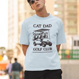 May include: A light blue t-shirt with a black graphic of cats riding in a golf cart. The text "Cat Dad Golf Club" is printed on the shirt.
