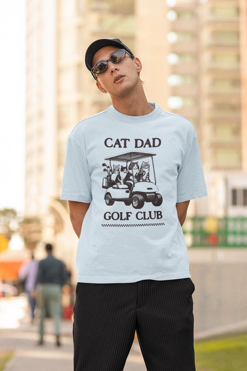 May include: A light blue t-shirt with a black graphic of cats riding in a golf cart. The text "Cat Dad Golf Club" is printed on the shirt.