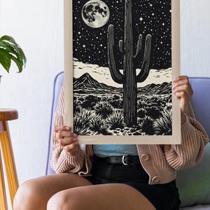 May include: A framed black and white print depicting a desert scene with a large cactus, mountains, and a starry night sky with a full moon. The artwork is held by a person.