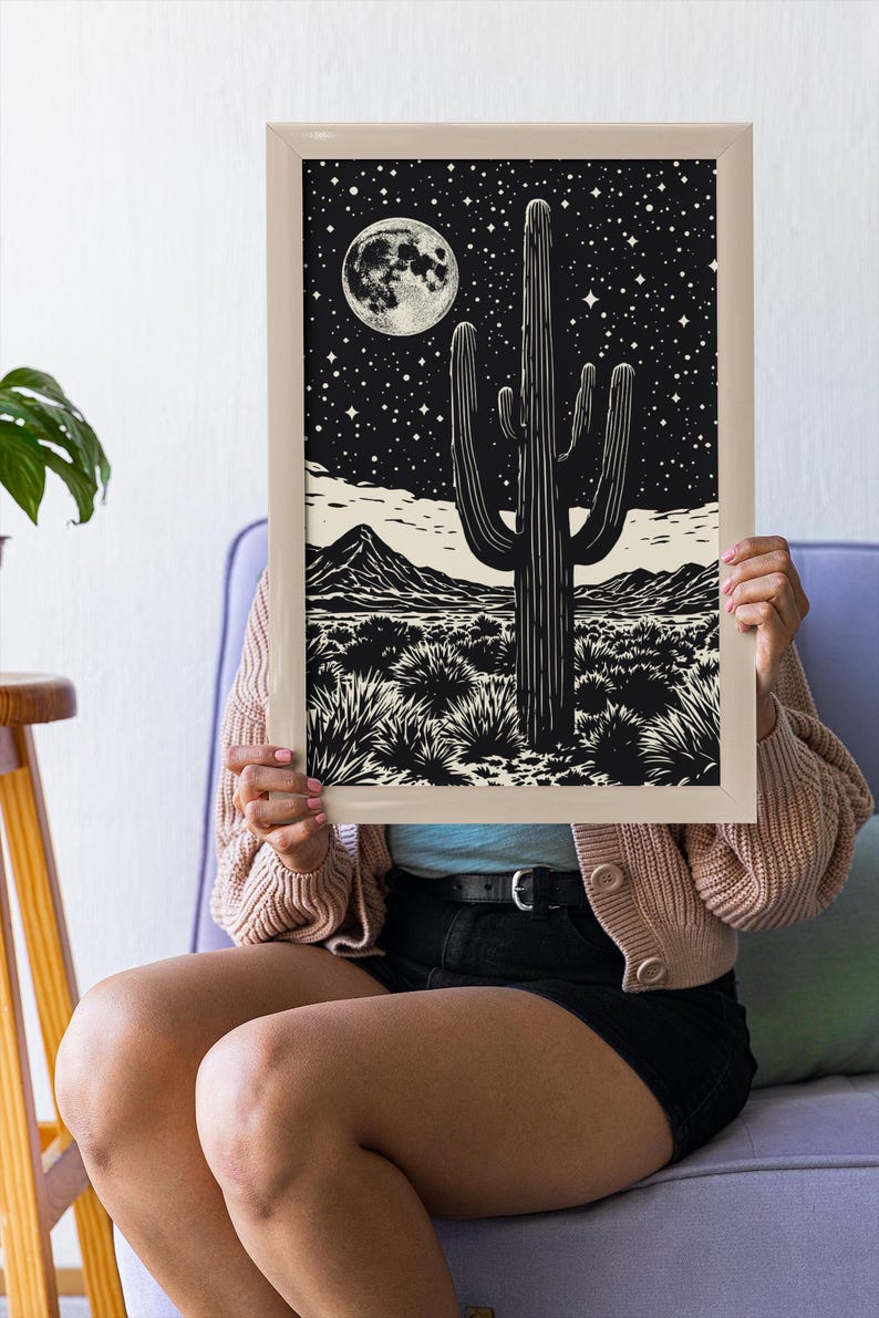 May include: A framed black and white print depicting a desert scene with a large cactus, mountains, and a starry night sky with a full moon. The artwork is held by a person.