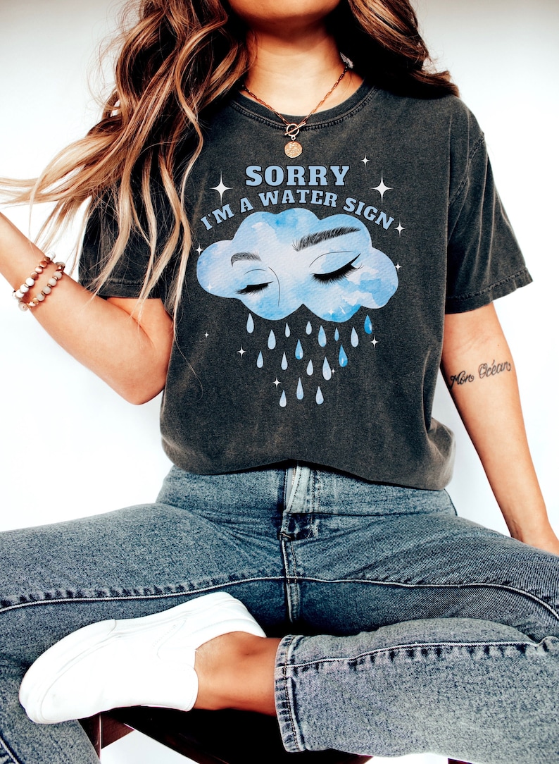 May include: A dark gray t-shirt with a blue watercolor cloud with eyes closed and raindrops falling. The text on the shirt reads "Sorry I'm a water sign".