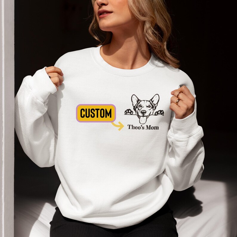 May include: White sweatshirt with a yellow rectangle that says "CUSTOM" and an arrow pointing to a black and white illustration of a dog peeking over a surface with the text "Theo's Mom" underneath.