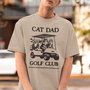 May include: A beige t-shirt with a graphic of three cats riding in a golf cart. The text "Cat Dad Golf Club" is printed on the shirt.