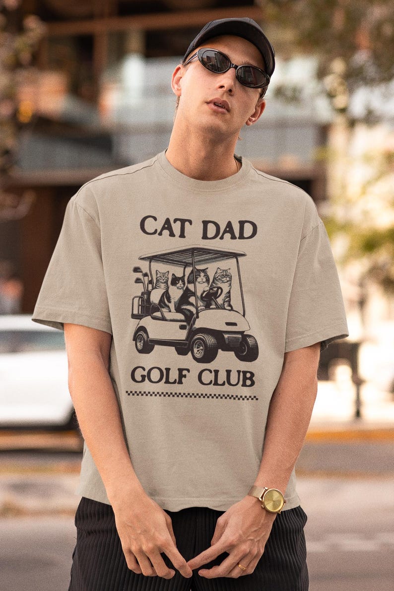 May include: A beige t-shirt with a graphic of three cats riding in a golf cart. The text "Cat Dad Golf Club" is printed on the shirt.