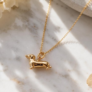 May include: A gold-coloured necklace with a dachshund pendant. The pendant is a detailed miniature sculpture of the dog breed. The necklace chain is delicate and also gold-coloured. The background is a marble surface with soft floral accents.
