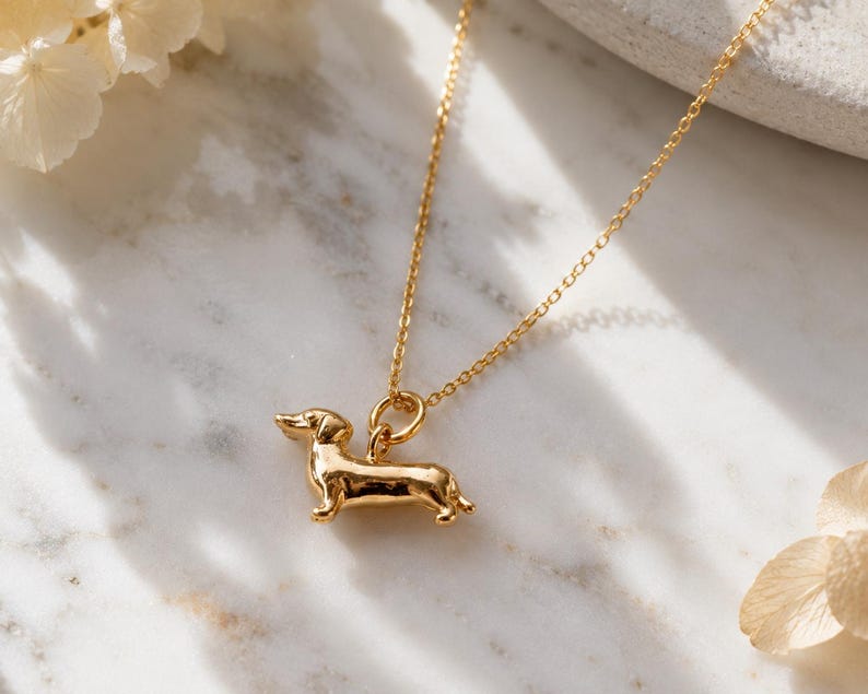 May include: A gold-coloured necklace with a dachshund pendant. The pendant is a detailed miniature sculpture of the dog breed. The necklace chain is delicate and also gold-coloured. The background is a marble surface with soft floral accents.