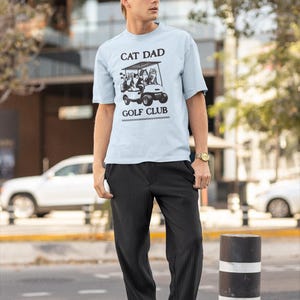 May include: A man wearing a light blue t-shirt with the text "Cat Dad Golf Club" and a graphic of cats in a golf cart. He is wearing black pants and black shoes. He is also wearing a black baseball cap and sunglasses.