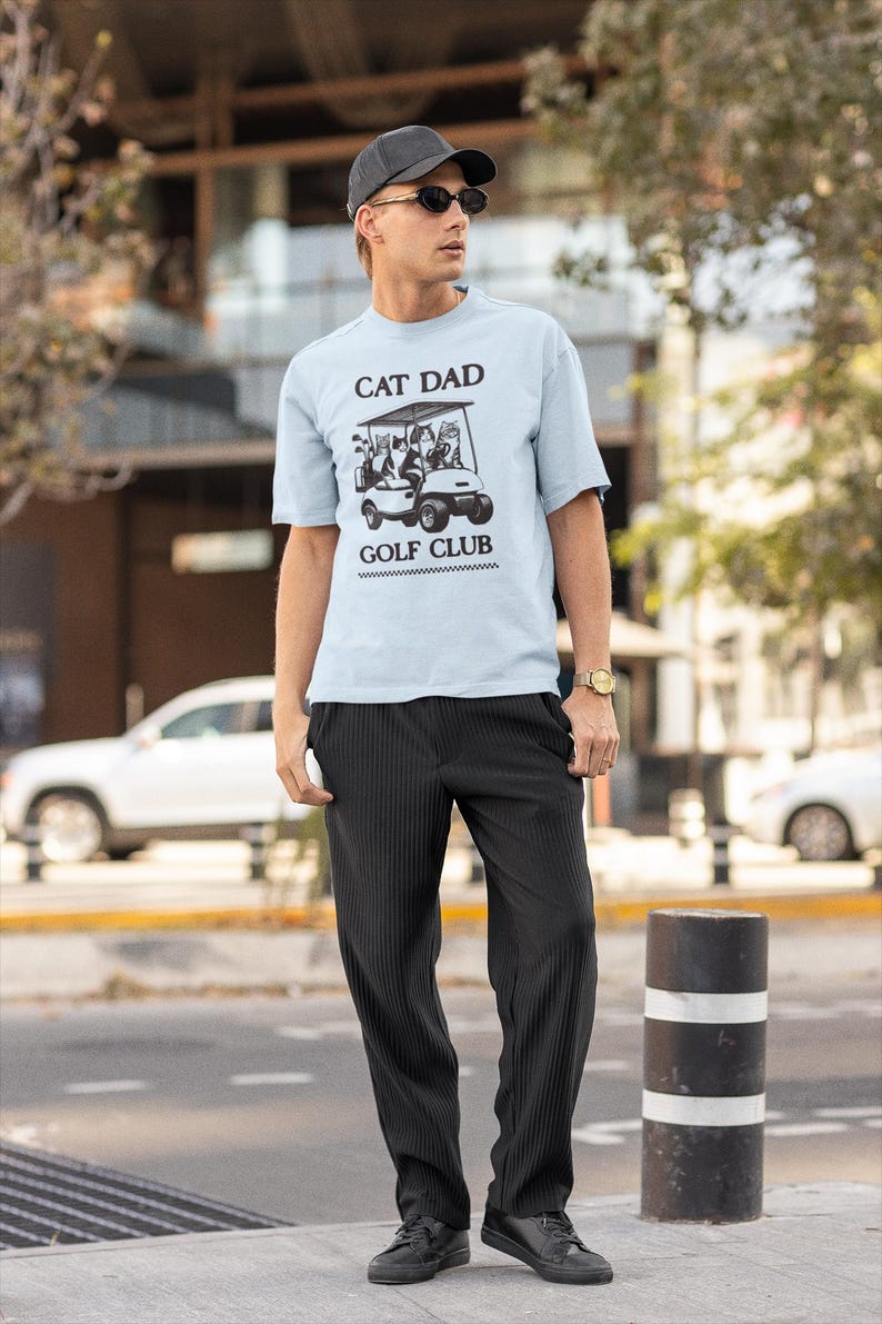May include: A man wearing a light blue t-shirt with the text "Cat Dad Golf Club" and a graphic of cats in a golf cart. He is wearing black pants and black shoes. He is also wearing a black baseball cap and sunglasses.