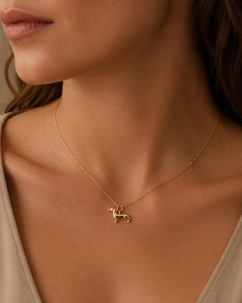 May include: A gold-coloured necklace with a dachshund pendant. The pendant is a detailed miniature of the dog breed. The necklace is worn around the neck, and the chain is delicate and thin.