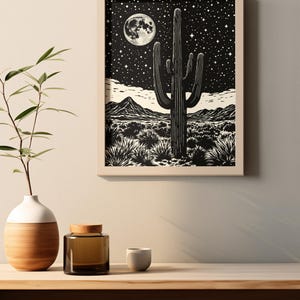 May include: Framed black and white print depicting a desert scene with a saguaro cactus under a starry night sky and a full moon. The artwork is displayed on a wooden shelf with a vase, a jar, and a small cup.