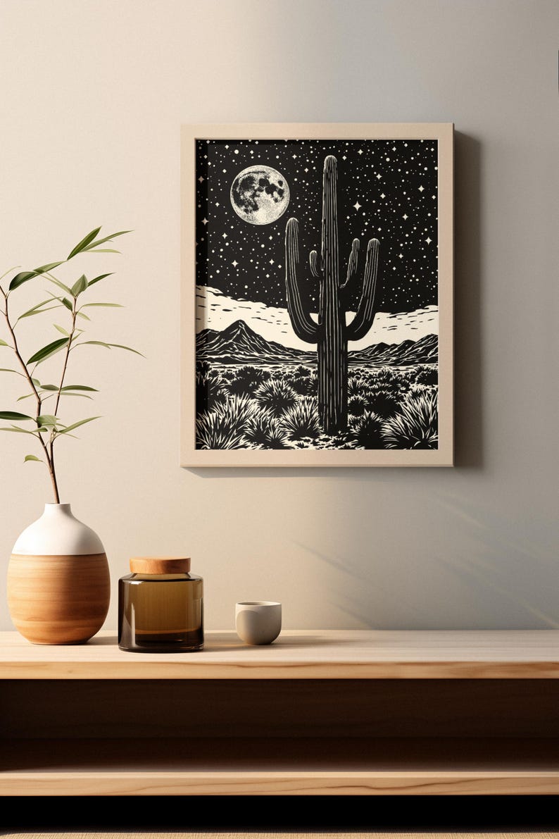 May include: Framed black and white print depicting a desert scene with a saguaro cactus under a starry night sky and a full moon. The artwork is displayed on a wooden shelf with a vase, a jar, and a small cup.