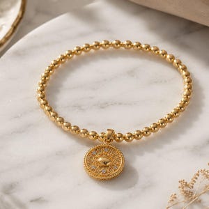 May include: A gold-colored beaded bracelet with a circular pendant. The pendant features a detailed design with small, sparkling accents. The bracelet is displayed on a white marble surface, with a neutral background.