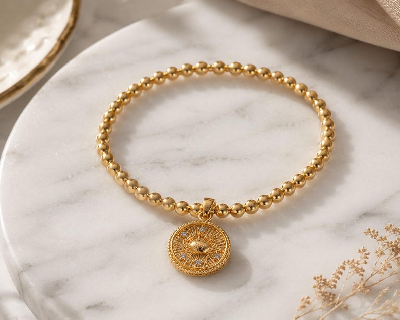 May include: A gold-colored beaded bracelet with a circular pendant. The pendant features a detailed design with small, sparkling accents. The bracelet is displayed on a white marble surface, with a neutral background.