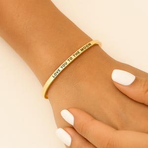May include: A gold-colored bangle bracelet with the inscription "LOVE YOU TO THE MOON" in a delicate, sans-serif font. The bracelet is worn on a wrist, showcasing its simple, elegant design. The background is a neutral, warm tone.