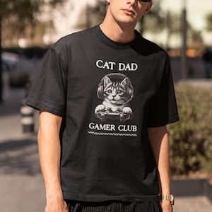 May include: Black t-shirt with the text "CAT DAD" above a graphic of a cat wearing headphones and holding a game controller. Below the graphic is the text "GAMER CLUB".