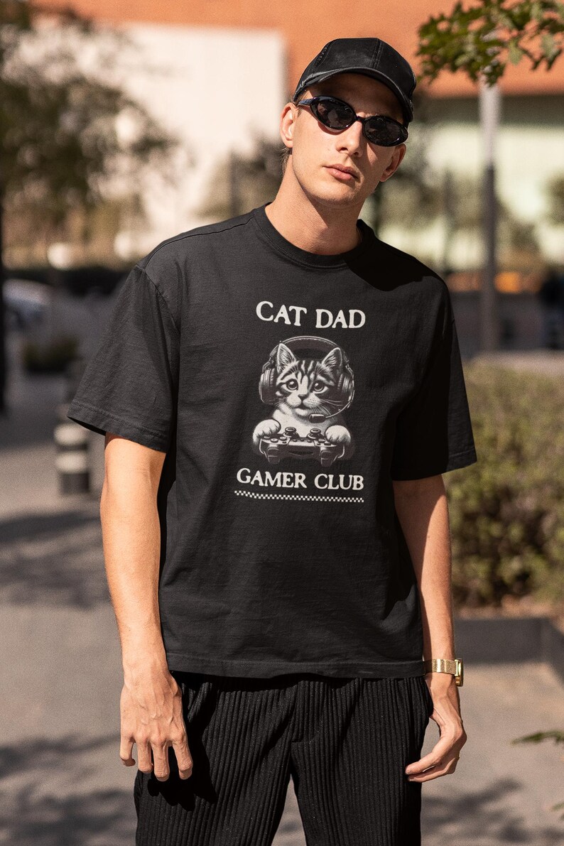May include: Black t-shirt with the text "CAT DAD" above a graphic of a cat wearing headphones and holding a game controller. Below the graphic is the text "GAMER CLUB".
