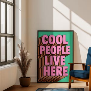 May include: A framed poster with the text "COOL PEOPLE LIVE HERE" in pink and orange, set against a green background. The poster is in a room with a blue chair, a decorative plant, and a blue rug.