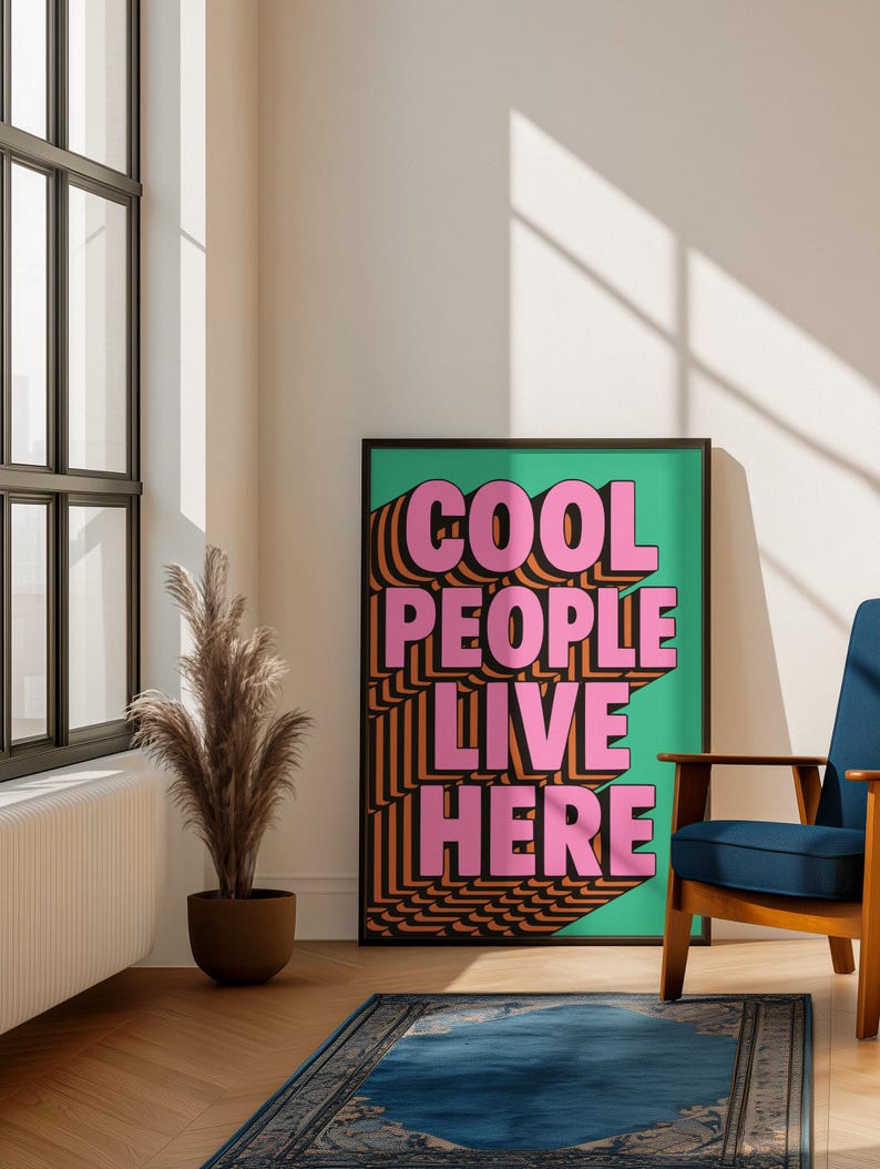May include: A framed poster with the text "COOL PEOPLE LIVE HERE" in pink and orange, set against a green background. The poster is in a room with a blue chair, a decorative plant, and a blue rug.