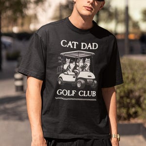 May include: Black t-shirt with a white graphic of cats riding in a golf cart. The text "CAT DAD GOLF CLUB" is printed on the shirt.