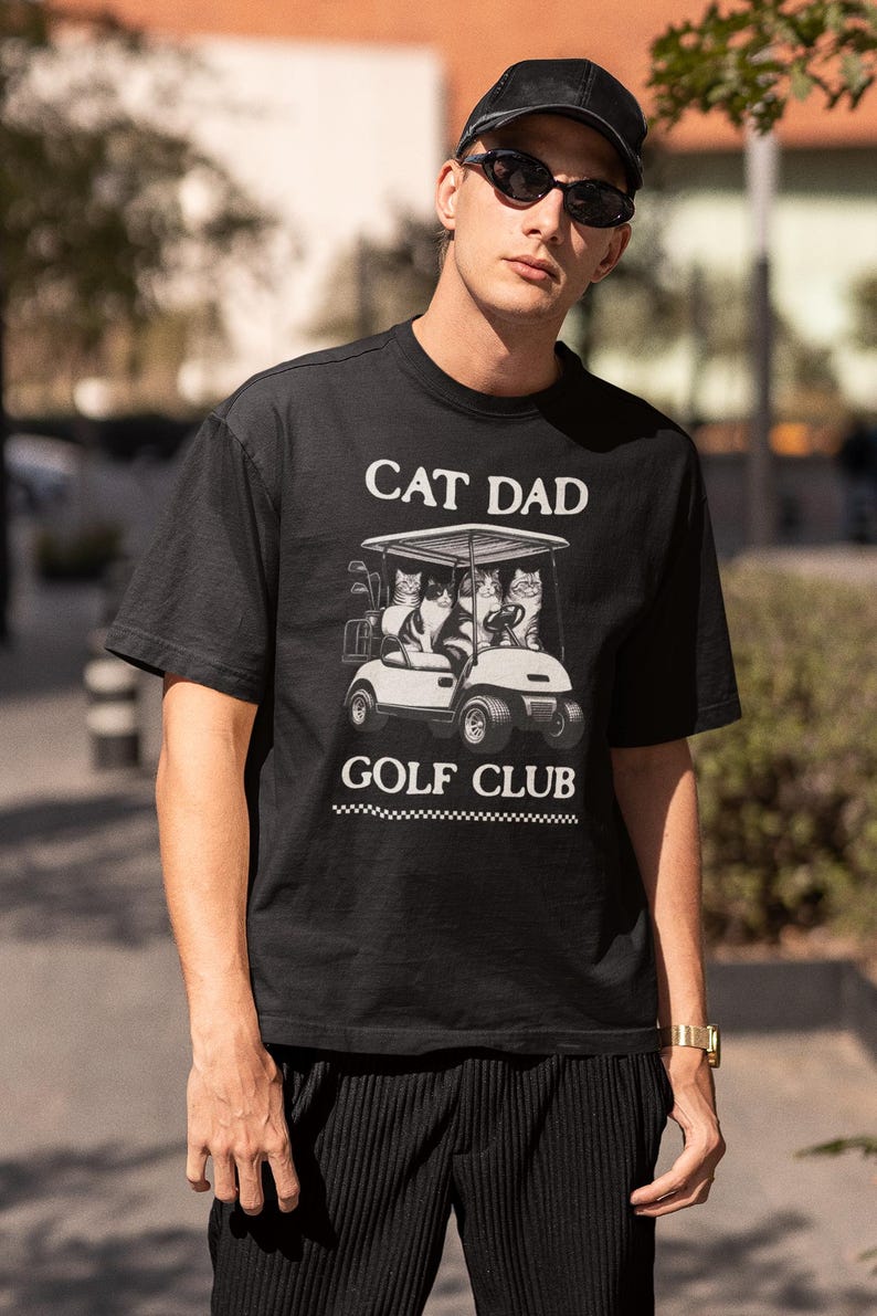 May include: Black t-shirt with a white graphic of cats riding in a golf cart. The text "CAT DAD GOLF CLUB" is printed on the shirt.
