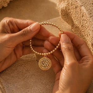 May include: Gold beaded bracelet with a round pendant. The pendant features a detailed design with small, sparkling accents. The bracelet is held against a neutral, sandy background, highlighting the jewelry's warm tones.