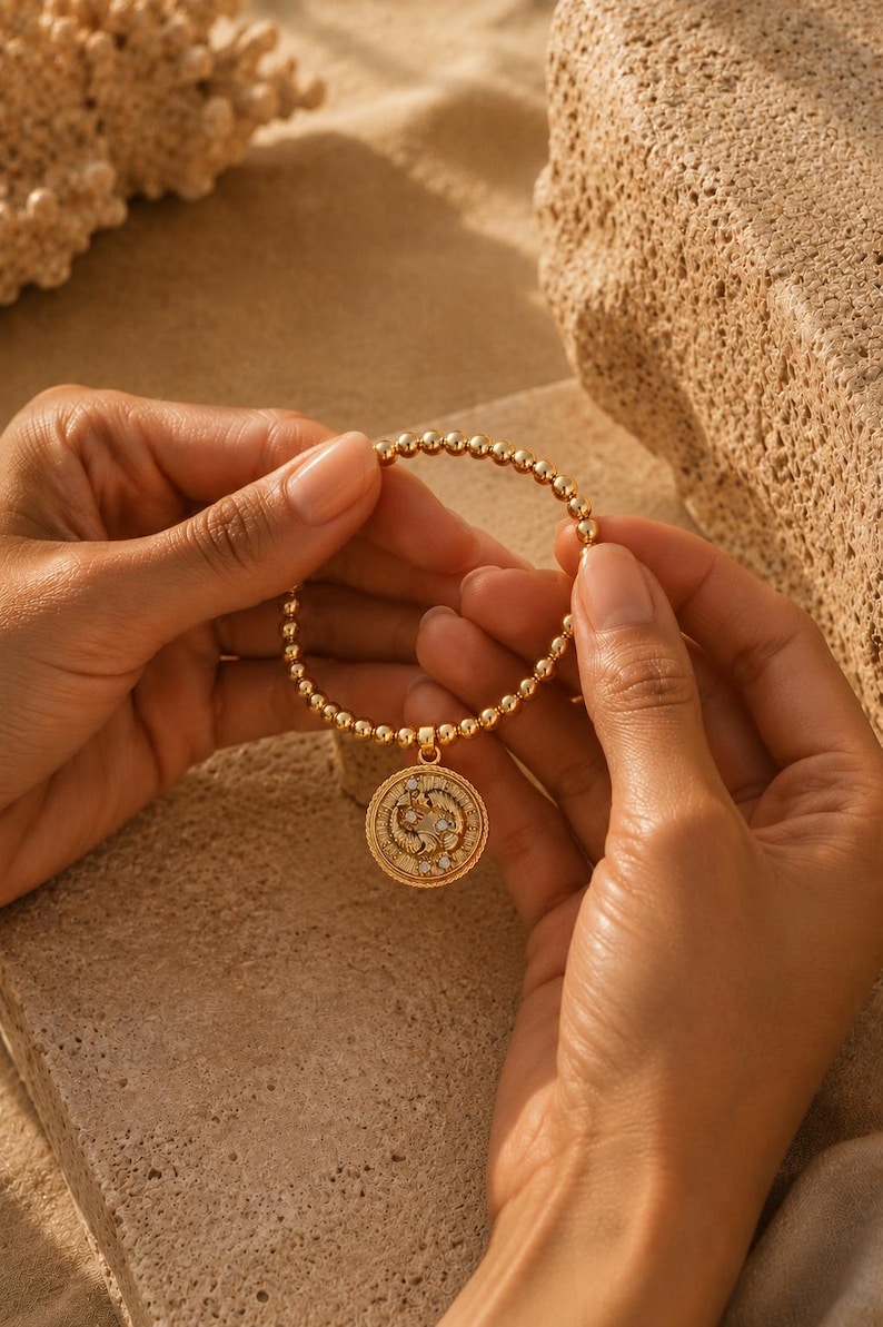 May include: Gold beaded bracelet with a round pendant. The pendant features a detailed design with small, sparkling accents. The bracelet is held against a neutral, sandy background, highlighting the jewelry's warm tones.
