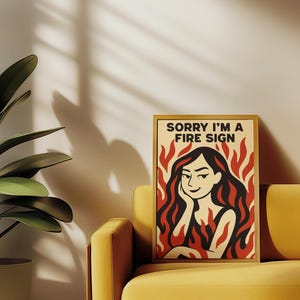 May include: A framed art print with the text "SORRY I'M A FIRE SIGN" features a woman with red hair and flames. The artwork is set against a cream background with a yellow sofa and a potted plant.