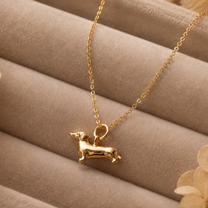 May include: A gold-coloured necklace with a dachshund pendant. The pendant is a detailed miniature sculpture of the dog breed. The necklace chain is delicate and hangs against a neutral background.