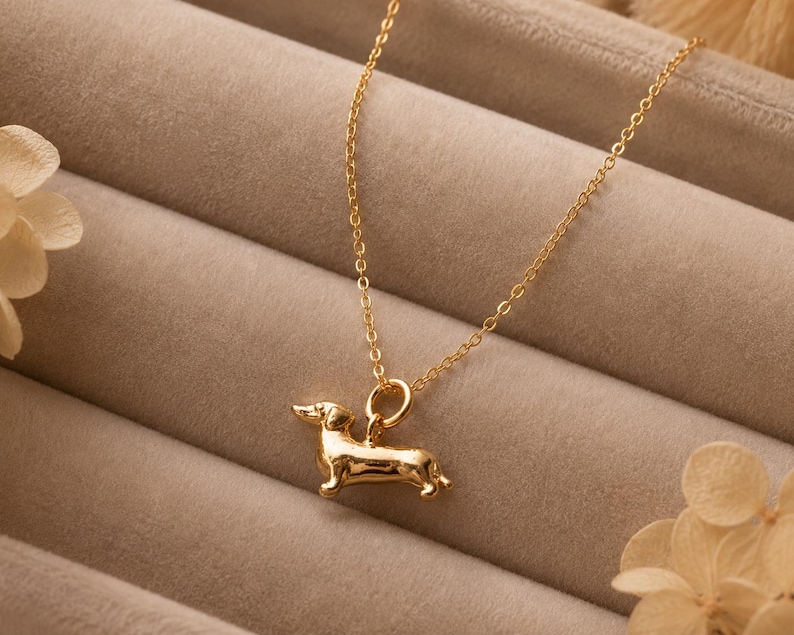 May include: A gold-coloured necklace with a dachshund pendant. The pendant is a detailed miniature sculpture of the dog breed. The necklace chain is delicate and hangs against a neutral background.