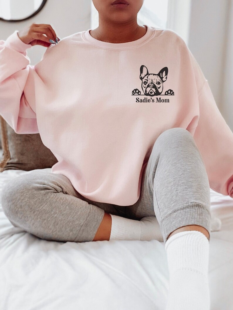 May include: A pink sweatshirt with a black graphic of a French bulldog peeking over the top and the text "Sadie's Mom" underneath.