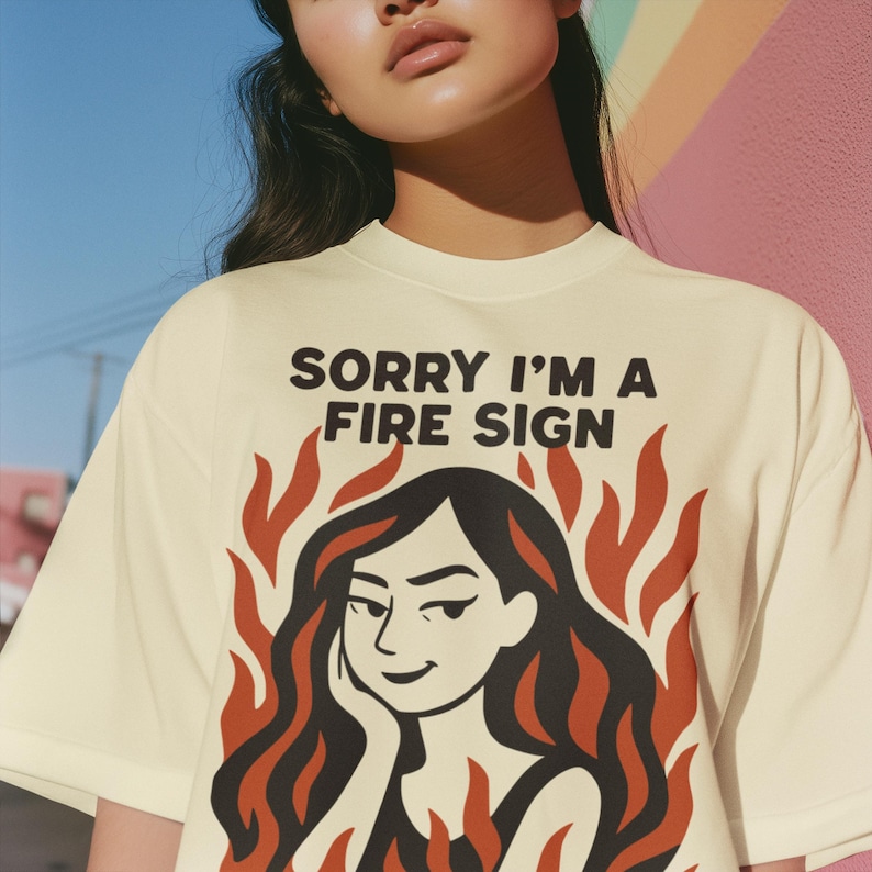 Puede incluir: Camiseta color crema con el texto "SORRY I'M A FIRE SIGN" en negro. El gr&aacute;fico presenta a una mujer con cabello negro y llamas rojas. La camiseta es de estilo casual, de manga corta.