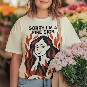 Puede incluir: Camiseta color crema con el texto "SORRY I'M A FIRE SIGN" sobre un gr&aacute;fico de una mujer con cabello negro y llamas rojas. La camiseta es de estilo casual, adecuada para el uso diario.